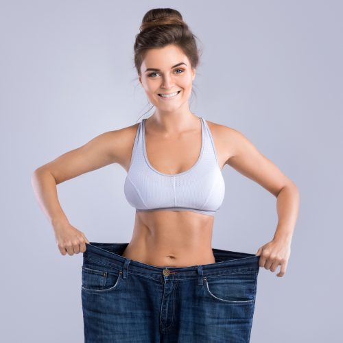 Happy woman wearing jeans after weight-loss on gray background